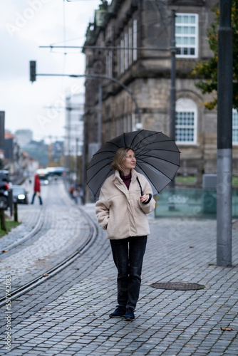 A woman with an umbrella stands on the sidewalk. © De Visu