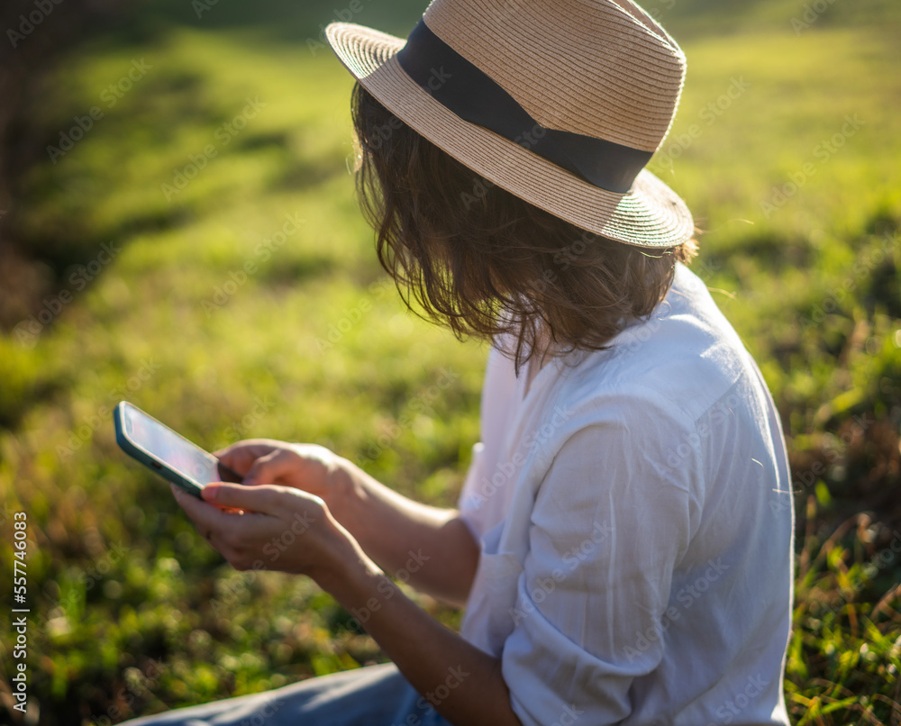 Fototapeta premium Happy pretty young cheerful woman in white shirt and sunglasses using smartphone while sitting on green grass on a sunny day