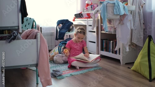 Little girl in pink dress reading book and eating apple while sitting on a rug in messy room at home photo