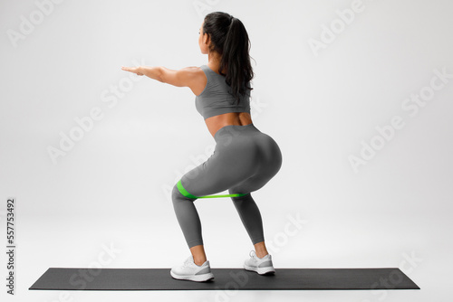 Athletic girl doing squats exercise for glute with resistance band on gray background. Fitness woman working out