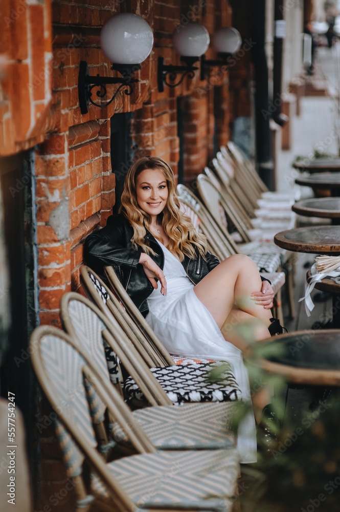 A beautiful curly smiling blonde bride, biker, lover of musical rock in a black leather jacket, white dress with a bouquet of reeds sits on chairs in the city on the street. wedding photography.