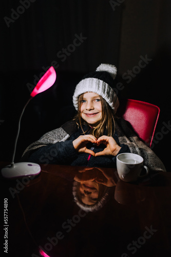 Blackout in Ukraine. A Ukrainian little girl, a child in warm clothes, a hat, a blanket, a blanket, sits at a table in a cold room in the dark with a lamp without light and shows a heart.