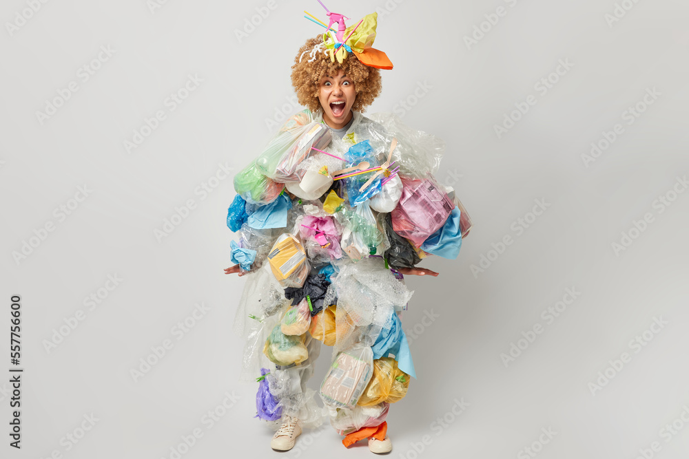 Full length shot of curly haired woman yells loudly made costume of plastic garbage takes care of environment isolated over grey background. Emotional female volunteer bothered by pollution. Full length shot of curly haired woman yells loudly made costume of plastic garbage takes care of environment isolated over grey background. Emotional female volunteer bothered by pollution.