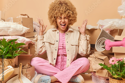 Quadro em tela Stressed curly haired woman gestures angrily screams loudly sits crossed legs surrounded by household stuff relocates to new apartment isolated over beige background