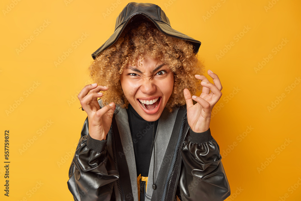 Photo of curly haired young woman screams with rage feels agressive expresses negativity gestures angrily wears black leather raincoat and hat isolated over yellow background tries to scare. Photo of curly haired young woman screams with rage feels agressive expresses negativity gestures angrily wears black leather raincoat and hat isolated over yellow background tries to scare.