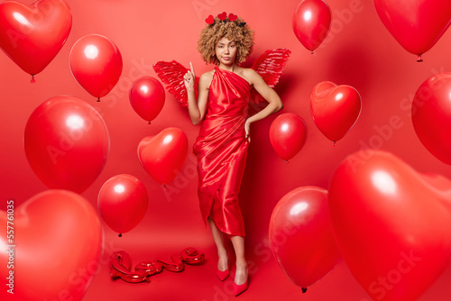 Full length shot of serious curly haired lady wears long dress fashionable shoes wings behind back indicates index finger above stands against bright red background with heart shaped balloons around