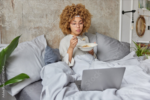 Serious woman eats oatmeal for breakfast relaxes in bed watches intersting film via laptop computer enjoys cozy morning at home wears spectacles and domestic clothes. Modern bedroom interior photo