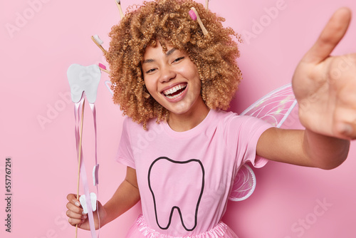 Horizontal shot of positive woman pretends being tooth fairy smiles broadly holds wand tells how to care about teeth smiles happily isolated over pink background. Female model comes on carnival party