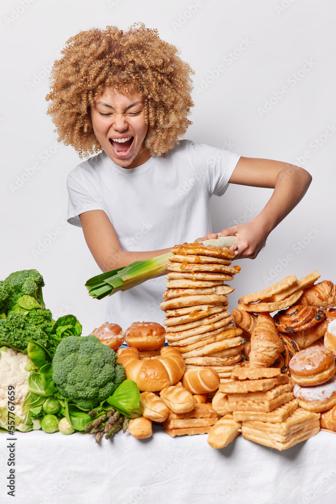 Vertical shot of unhappy emotional woman fights delicious dessert with leek surrounded by healthy and unhealthy food tries to keep to diet avoids eating yummy sweet desserts wears casual t shirt Vertical shot of unhappy emotional woman fights delicious dessert with leek surrounded by healthy and unhealthy food tries to keep to diet avoids eating yummy sweet desserts wears casual t shirt