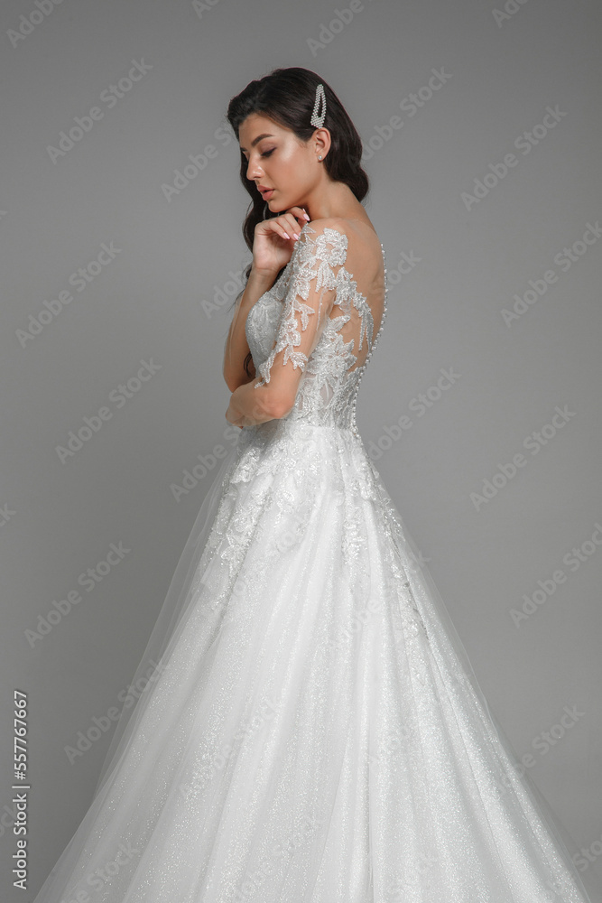 Side view of the young caucasian brunette bride in the beautiful white wedding dress with pearls and embroidery standing, posing with hands in the grey studio.