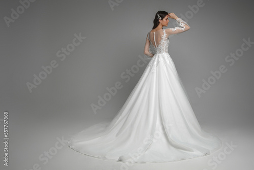 Back view of the beautiful brunette bride wearing modern white wedding dress with pattern and pearls on the back. Gorgeous female posing with hands on a grey isolated background.