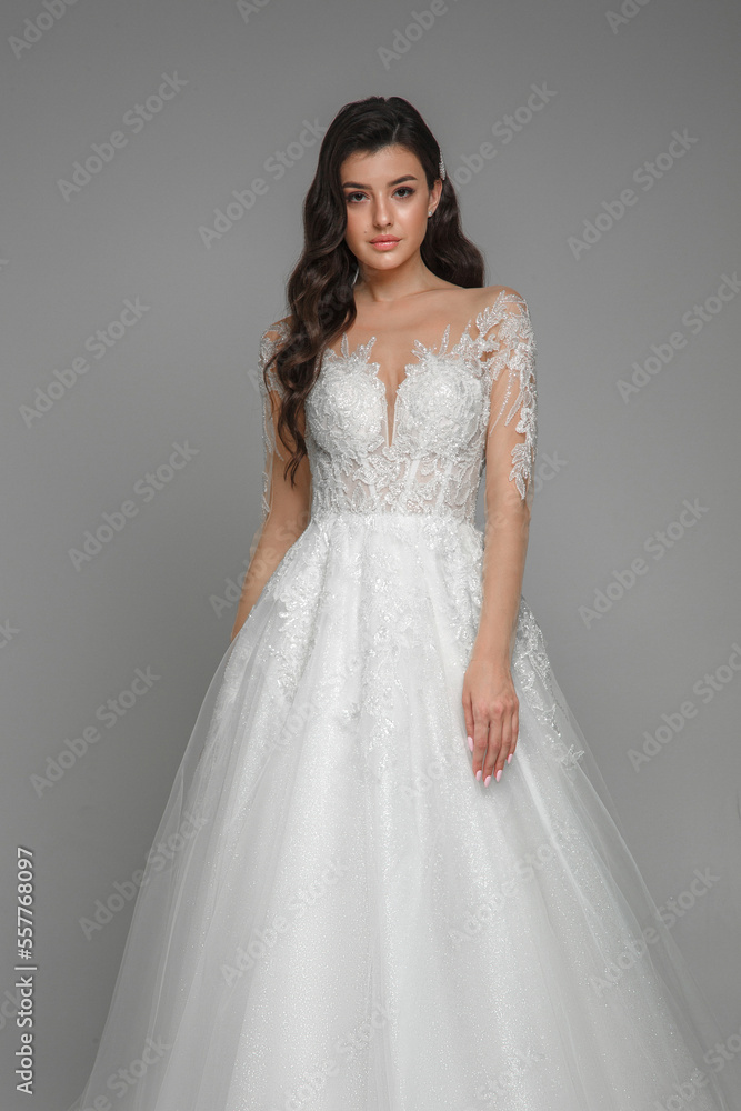 Portrait of the elegant beautiful woman wearing beautiful white wedding dress with embroidery on the sleeves. Attractive bride standing and posing. Gorgeous modern female on a grey background. Portrait of the elegant beautiful woman wearing beautiful white wedding dress with embroidery on the sleeves. Attractive bride standing and posing. Gorgeous modern female on a grey background.