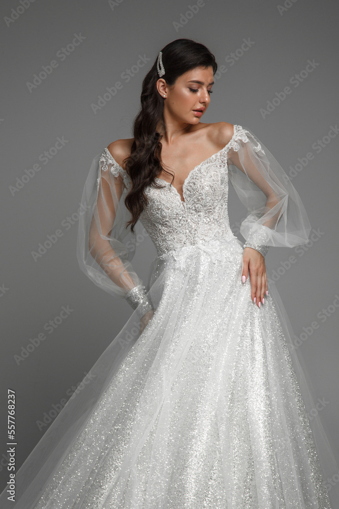 Brunette bride in the white wedding dress posing, holding hands on the waist. Gorgeous female on a grey isolated background. Brunette bride in the white wedding dress posing, holding hands on the waist. Gorgeous female on a grey isolated background.
