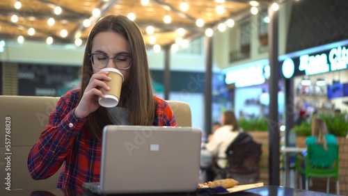 A young woman is working on a laptop in a cafe. A girl in a shirt and glasses sits with a laptop, eats pizza and drinks coffee.