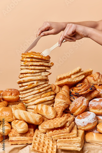 Unknown woman cuts delicious pancakes with jam with knife and fork against beige background. Variety of delicious dessets on table. Big assortment of appetizing sweet food. Unhealthy nutrition concept photo