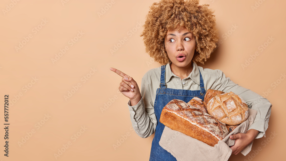 Impressed shocked woman with curly hair stands stupefied points index finger on blank space poses with delicious crusty bread shows something amazing wears denim apron isolated over brown background Impressed shocked woman with curly hair stands stupefied points index finger on blank space poses with delicious crusty bread shows something amazing wears denim apron isolated over brown background