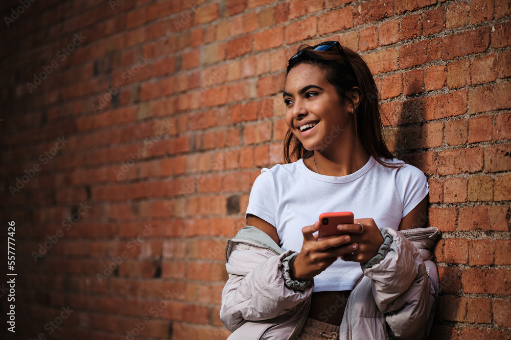 Young woman smiling while holding a mobile phone leaning against a brick wall.