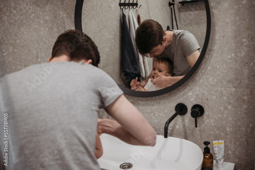  A father and son brushing their teeth together in a bathroom together while looking at the reflection.