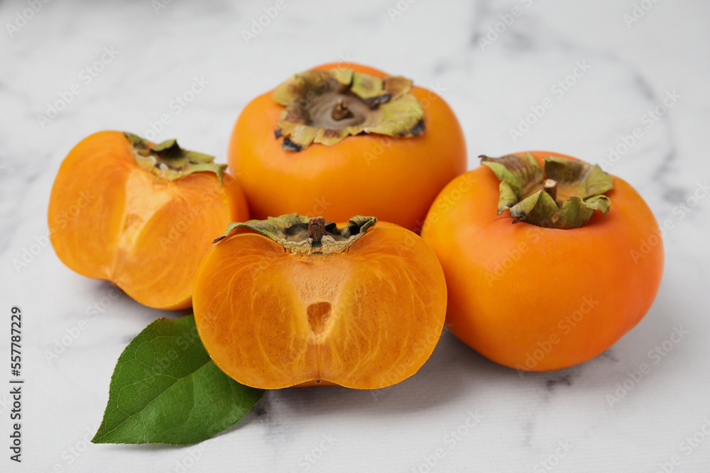 Whole and cut delicious ripe juicy persimmons with green leaf on white marble table Whole and cut delicious ripe juicy persimmons with green leaf on white marble table