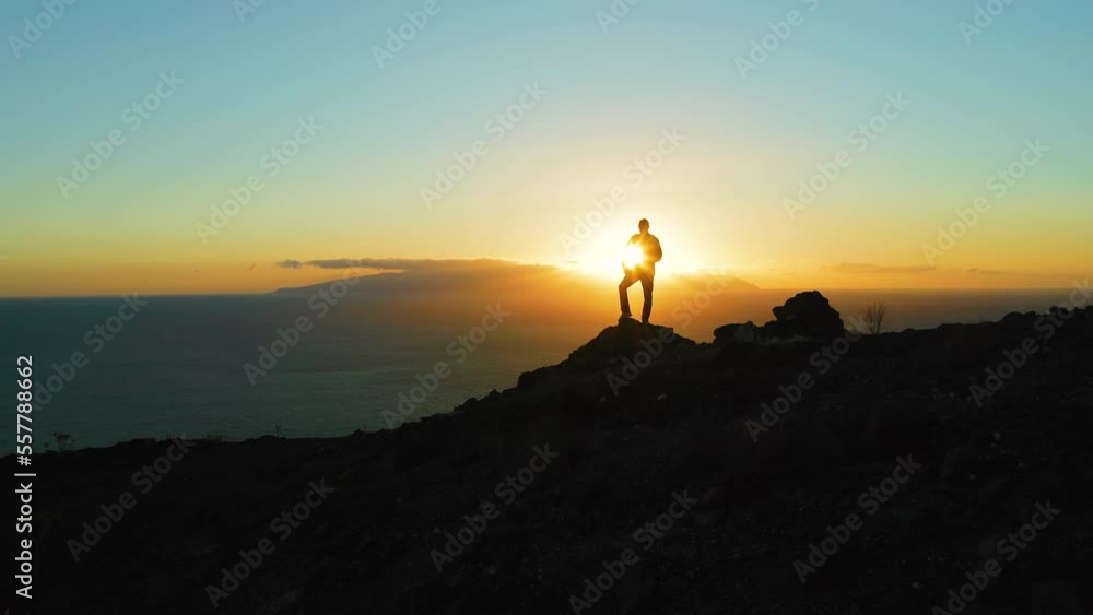 Hiking man in mountains watching sunset and horizon over beautiful ocean landscape. Male hiker silhouette against cloudy colorful sky with sunburst, sun flare. Hiking man in mountains watching sunset and horizon over beautiful ocean landscape. Male hiker silhouette against cloudy colorful sky with sunburst, sun flare.