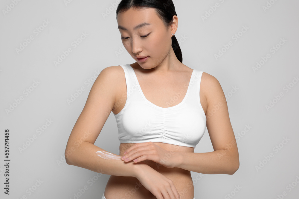 Beautiful young Asian woman applying body cream onto arm on light grey background Beautiful young Asian woman applying body cream onto arm on light grey background