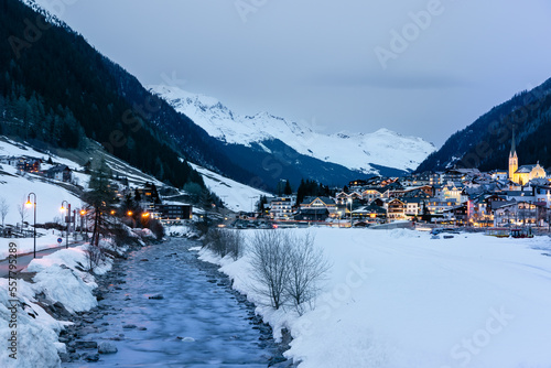 Winter scene in Ischgl ski resort in Austria with river flowing into distance and snowcapped mountains in background and illuminated village lit up as nightfall arrives