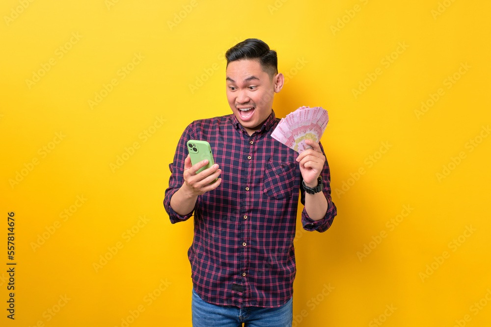 Surprised young Asian man using mobile phone and holding money banknotes isolated over yellow background. Fast credits concept Surprised young Asian man using mobile phone and holding money banknotes isolated over yellow background. Fast credits concept