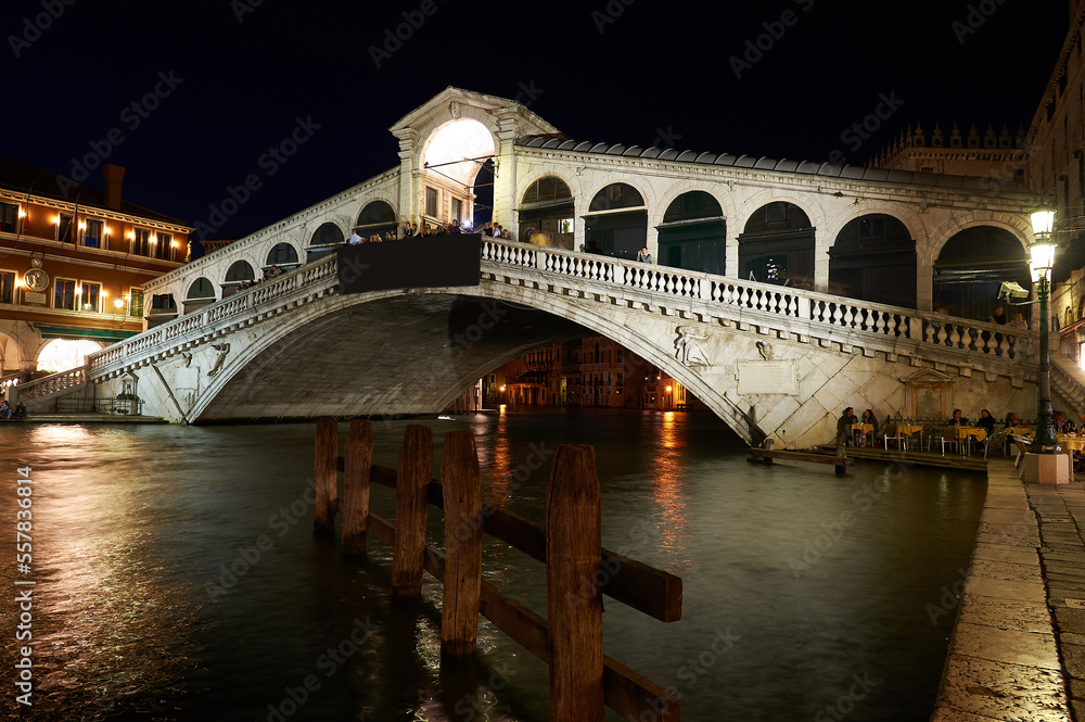 Night view of the Venetian Grand Canal with the Rialto Bridge in the background with people having dinner under the bridge in the moonlight