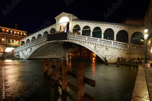 Night view of the Venetian Grand Canal with the Rialto Bridge in the background with people having dinner under the bridge in the moonlight