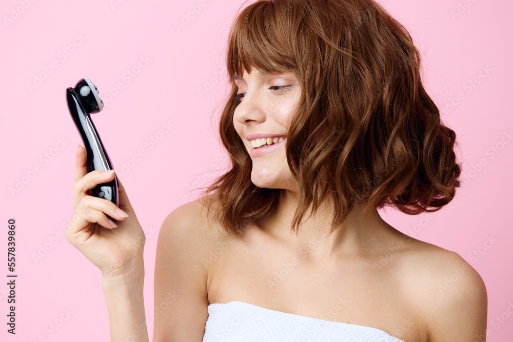 a happy, joyful, young woman stands with beautifully styled hair on a pink background and holding a black electric massager in her hand, looks at him with admiration a happy, joyful, young woman stands with beautifully styled hair on a pink background and holding a black electric massager in her hand, looks at him with admiration