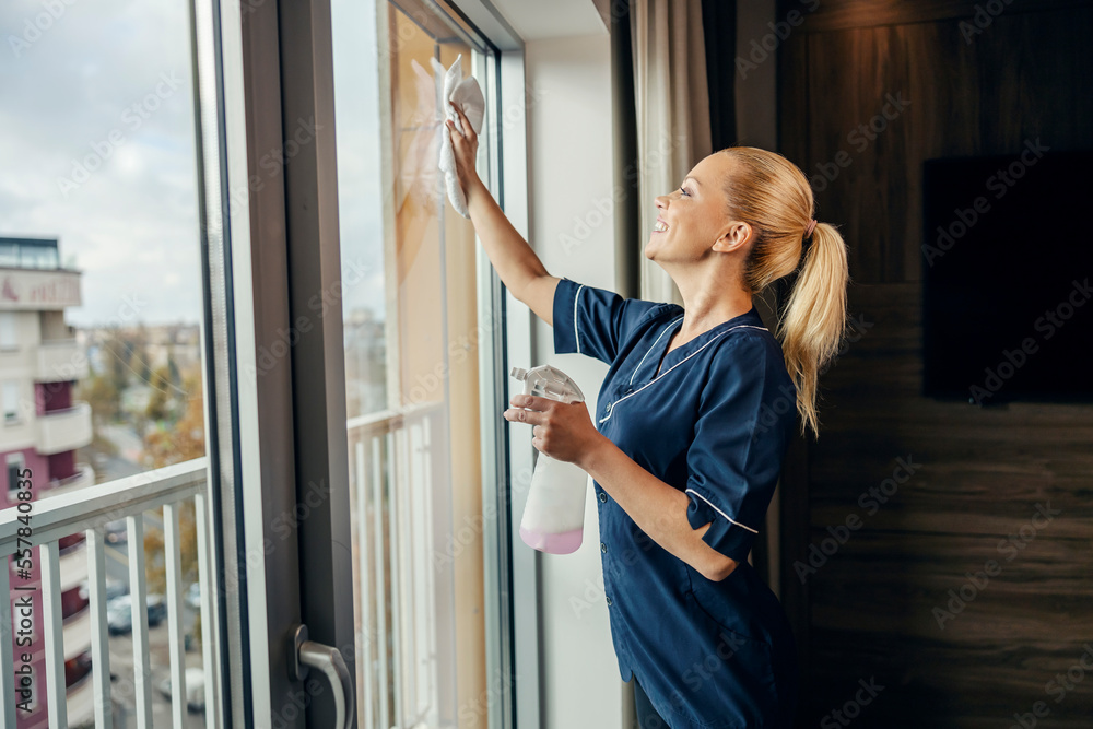 A happy maid is cleaning windows in hotel room with detergent and cloth. - obrazy, fototapety, plakaty 