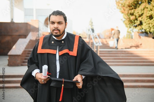 Portrait of handsome indian graduate student in graduation glow with diploma.