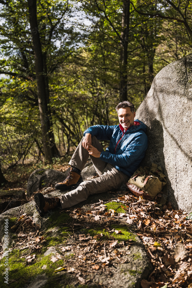 Smiling man with backpack sitting by rock in forest Smiling man with backpack sitting by rock in forest
