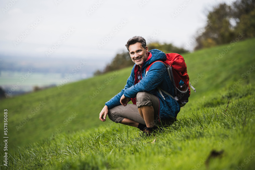 Happy mature man with backpack crouching on meadow Happy mature man with backpack crouching on meadow