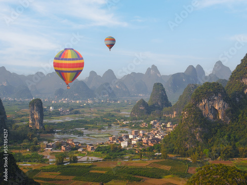 High angle view of hot air balloon flying over pretty rural village in countryside of Yangshuo county, China. Aerial view of mountains of limestone formations; rice fields in foreground.