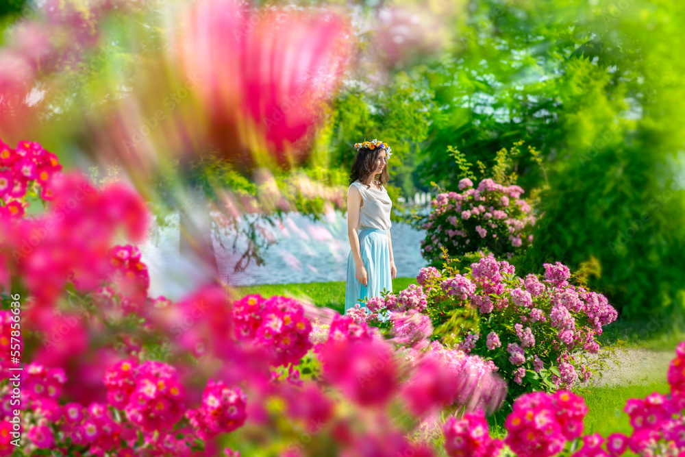 Outdoor portrait of young happy smiling fantasy woman walking among flowers. Young beautiful goddess enjoys spring nature in bright sunlight. Fairy tale image art photo with pink roses frame