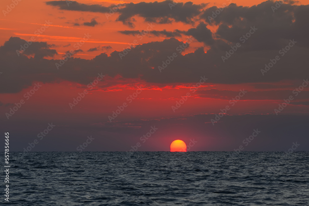 The very first moments of the sunrise, when sun rises above the sea. Sunrise over the ocean landscape with big red sun, orange sky, fluffy clouds, dark blue water. Spiritual new day natural background