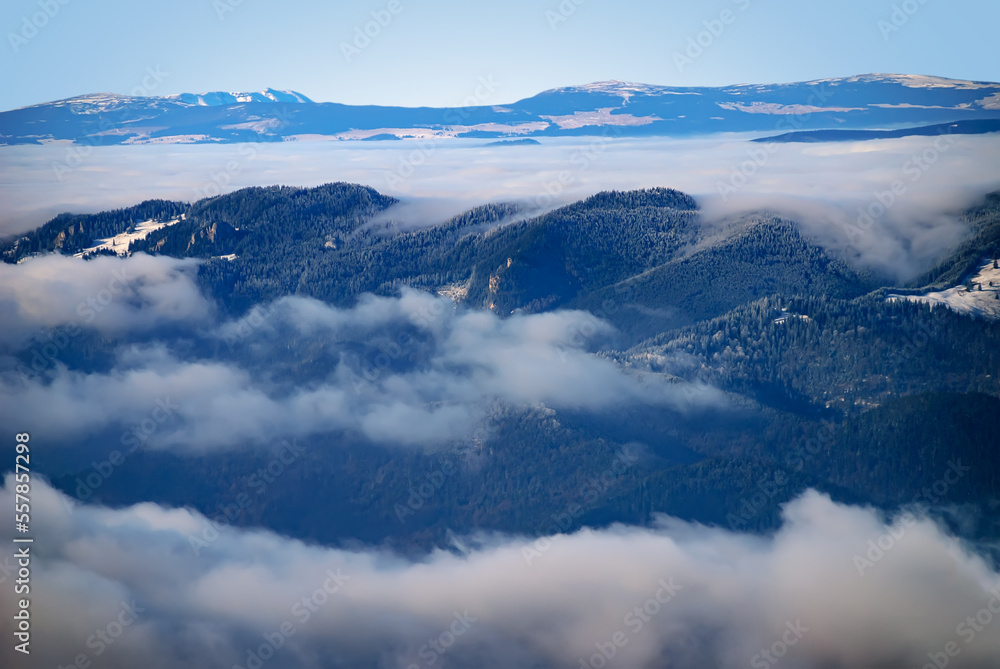 An epic ocean of clouds and fog in the winter mountains landscape, aerial view. Huge white clouds come in waves over the foggy valleys, mountain peaks rise over like islands. Dramatic overcast sky