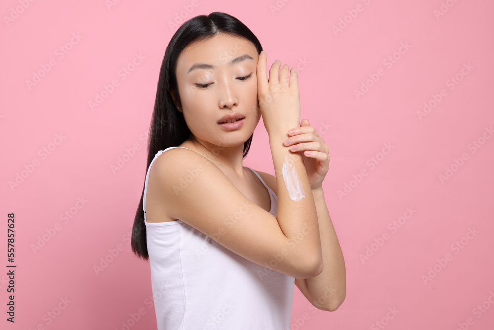 Beautiful young Asian woman applying body cream onto arm on pink background Beautiful young Asian woman applying body cream onto arm on pink background