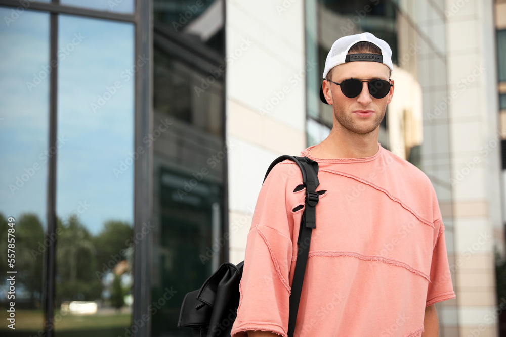 Handsome young man with stylish sunglasses and backpack near building outdoors, space for text Handsome young man with stylish sunglasses and backpack near building outdoors, space for text