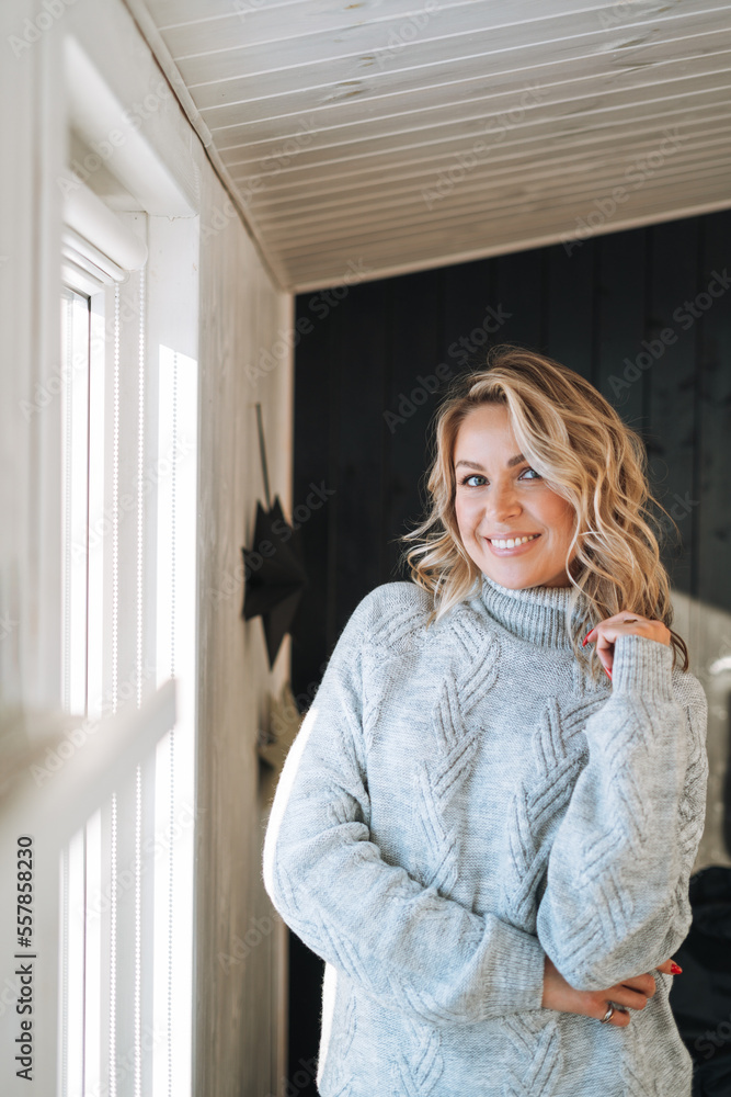 Smiling woman forty year with blonde curly hair in grey knitted sweater near window at house Smiling woman forty year with blonde curly hair in grey knitted sweater near window at house