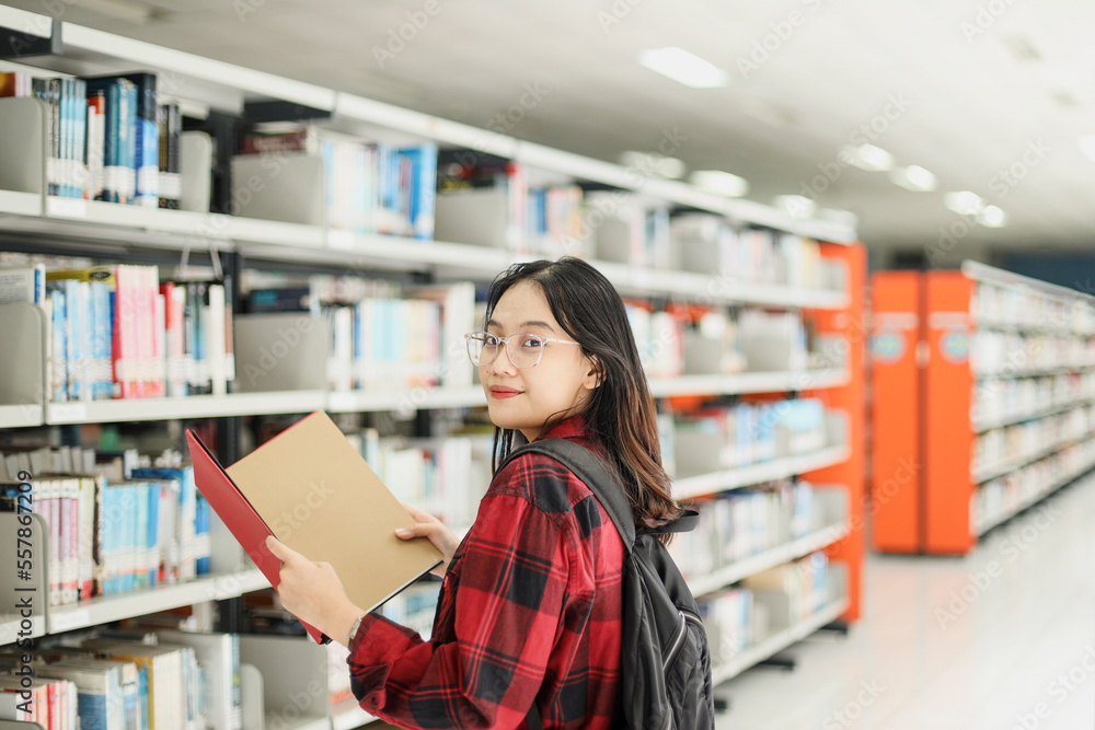 Female student in casual style is opening books and smiling to the camera against blurred library background. Female student in casual style is opening books and smiling to the camera against blurred library background.