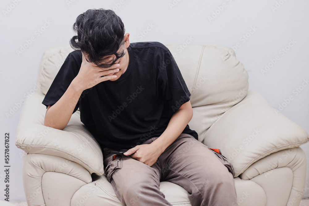 Unpleasant pain. Sad unhappy young man sitting on the sofa and holding his forehead feeling stress and frustrated.