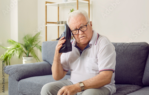 Senior man with dementia using everyday objects in an incorrect way. Retired old man with Alzheimer's disease sitting on the couch and holding a shoe at his ear as if talking on the telephone photo