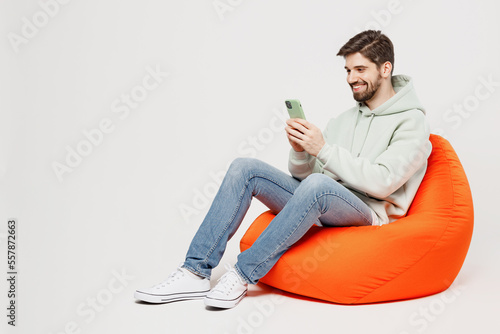 Full body young satisfied happy fun cheerful man wear mint hoody sit in bag chair hold in hand use mobile cell phone isolated on plain solid white background studio portrait. People lifestyle concept.