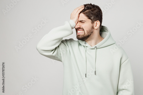 Young displeased sad unshaven caucasian man wear mint hoody put hand on face facepalm epic fail mistaken omg gesture isolated on plain solid white background studio portrait. People lifestyle concept.