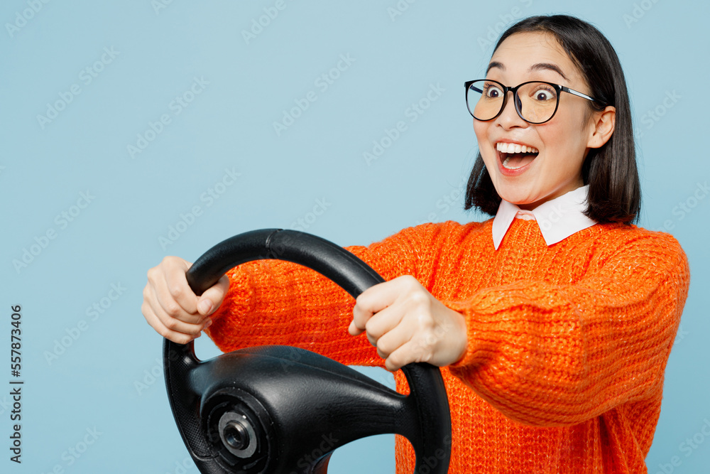 Side view young excited happy woman of Asian ethnicity wear orange sweater glasses hold steering wheel driving car isolated on plain pastel light blue cyan background studio. People lifestyle concept. Side view young excited happy woman of Asian ethnicity wear orange sweater glasses hold steering wheel driving car isolated on plain pastel light blue cyan background studio. People lifestyle concept.