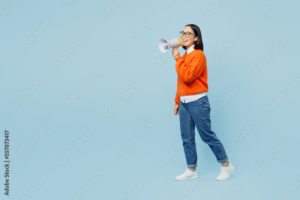 Full body shocked excited happy young woman of Asian ethnicity wear orange sweater glasses hold megaphone scream announces discounts sale Hurry up isolated on plain pastel light blue cyan background. Full body shocked excited happy young woman of Asian ethnicity wear orange sweater glasses hold megaphone scream announces discounts sale Hurry up isolated on plain pastel light blue cyan background.