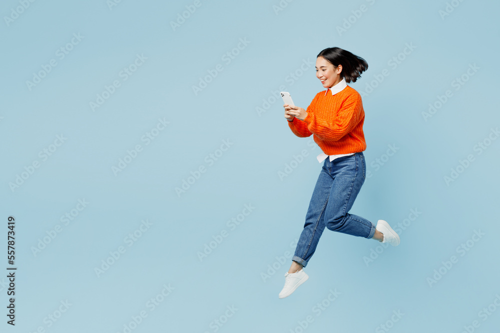 Full body young woman of Asian ethnicity wear orange sweater glasses jump high hold in hand use mobile cell phone isolated on plain pastel light blue cyan background studio. People lifestyle concept. Full body young woman of Asian ethnicity wear orange sweater glasses jump high hold in hand use mobile cell phone isolated on plain pastel light blue cyan background studio. People lifestyle concept.