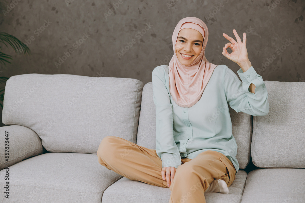 Young muslim woman in hijab casual clothes showing okay ok gesture sits on sofa couch stay at